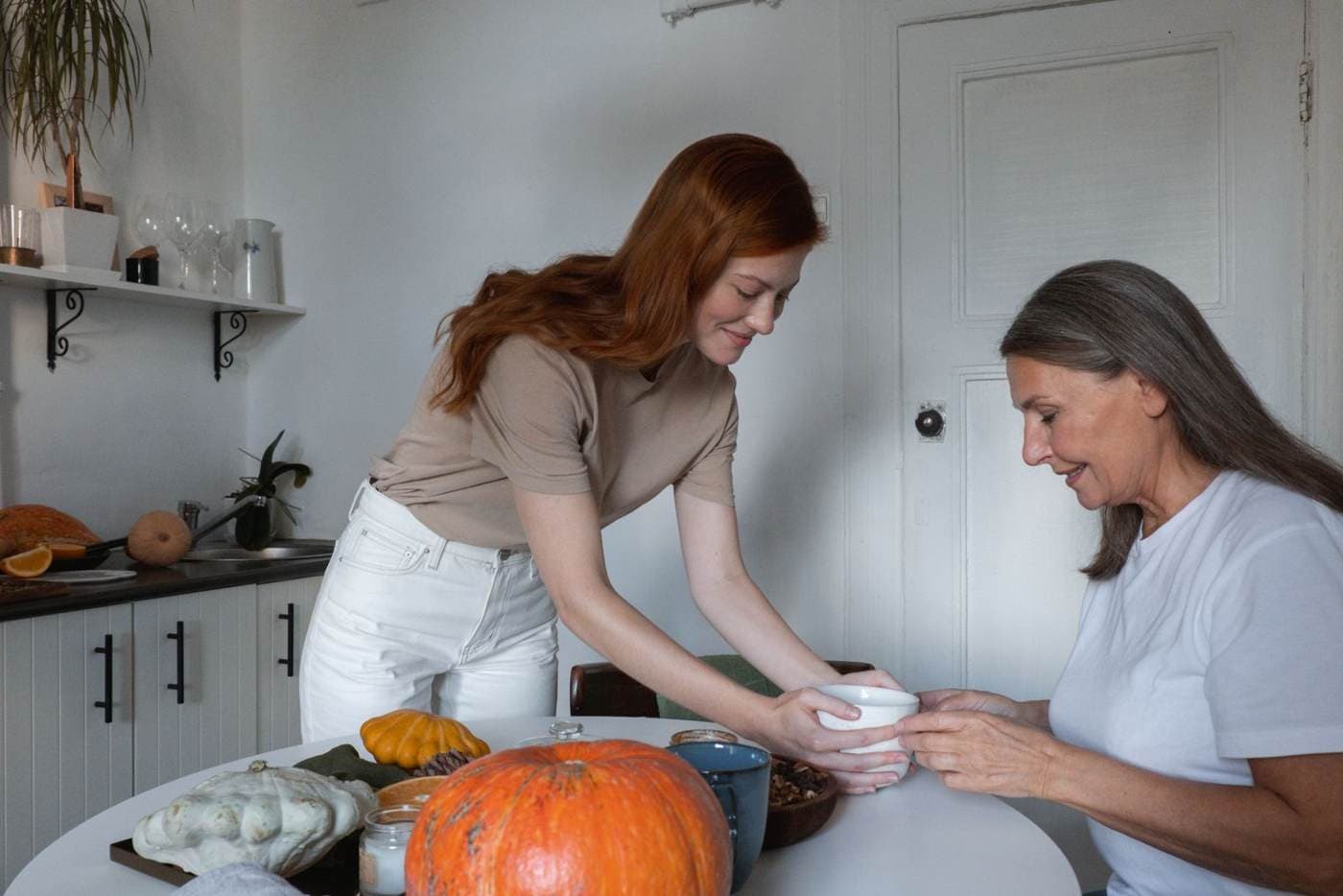 Una hija adulta está de pie y le pasa una taza, al otro lado de la mesa, a su madre mayor sentada. Sobre la mesa hay calabazas pequeñas.