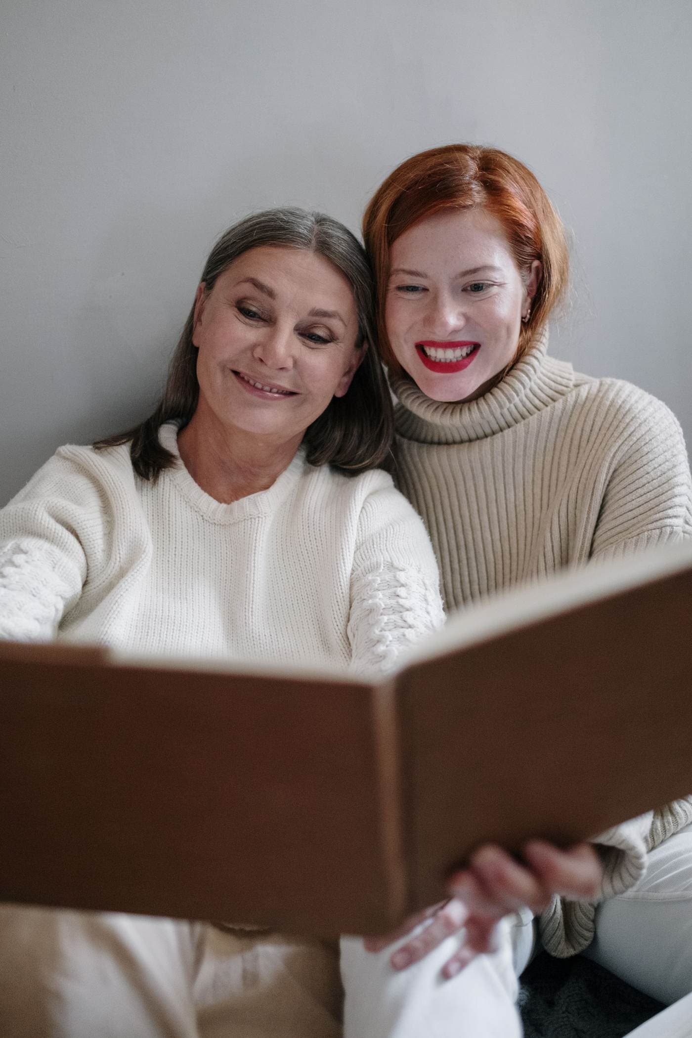 Una mujer mayor con el pelo hasta los hombros, entre gris y castaño, y una mujer más joven pelirroja están sentadas juntas en un interior y sonríen mientras miran un libro abierto que sostienen juntas.