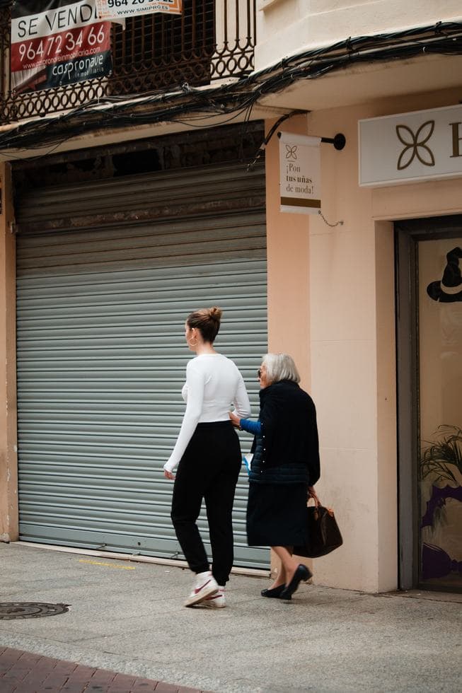 Una mujer más joven, vista de espaldas, camina por una acera mientras una mujer mayor a su lado le apoya una mano en el brazo, frente a una tienda con un cartel de se vende.