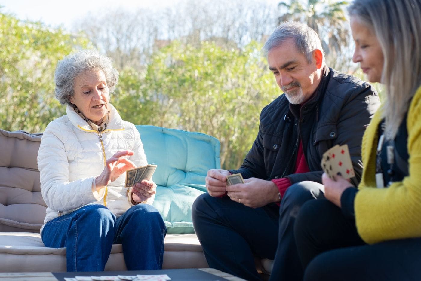 Three people play cards outdoors: an older woman and a gray-haired man look down at their cards while a blonde woman sits in profile.