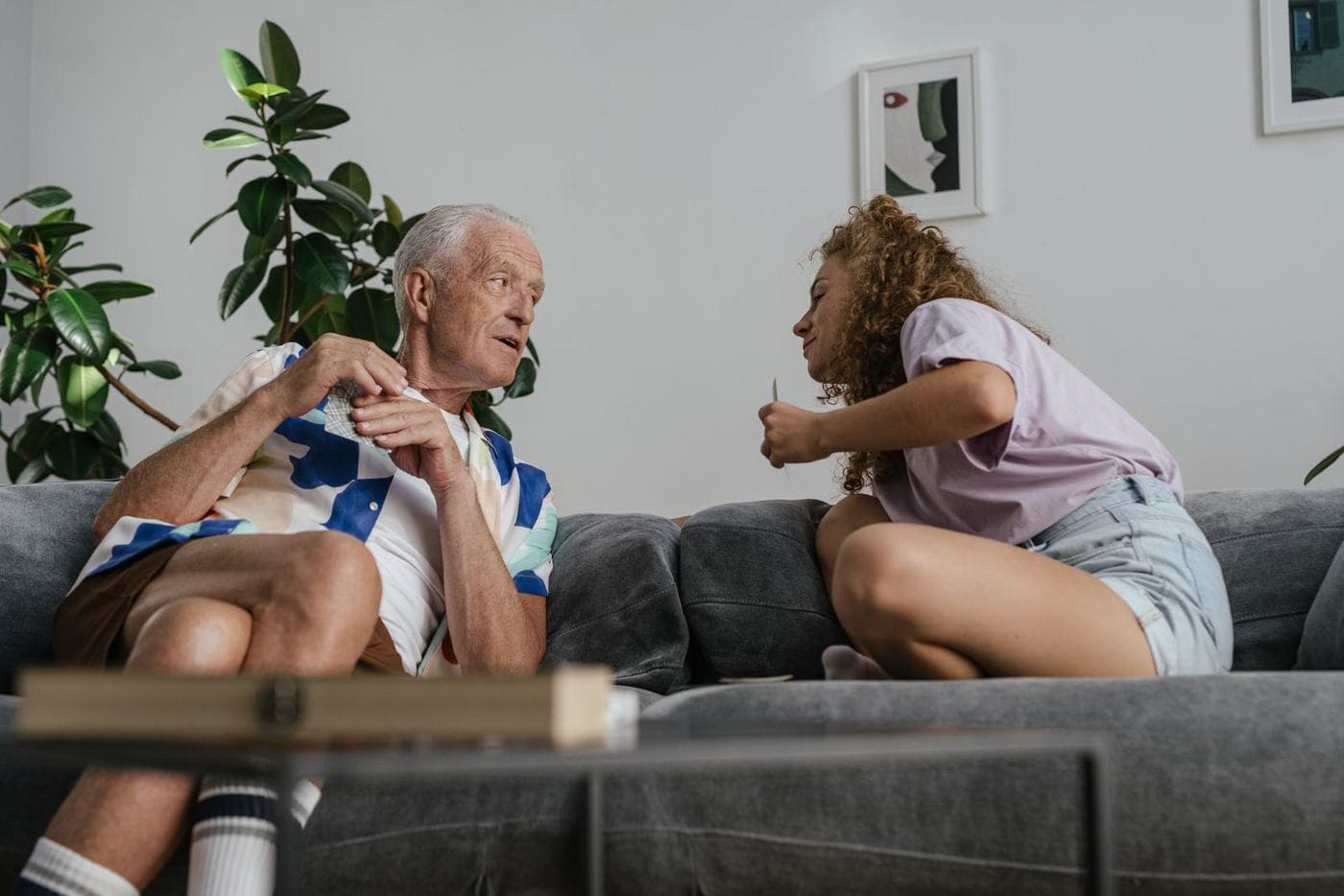 An older man and a younger woman or teenager sit facing each other on a gray sofa at home.